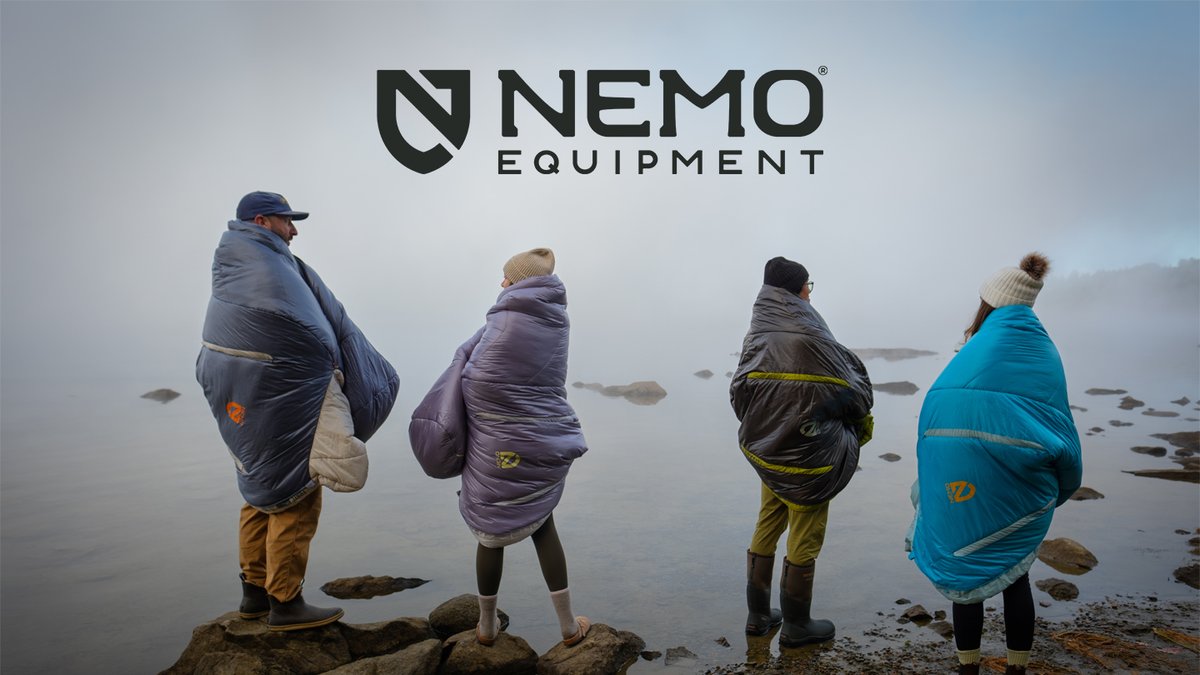 Four people wrapped in colorful Nemo sleeping bags stand on a rocky shore looking out at a misty lake.