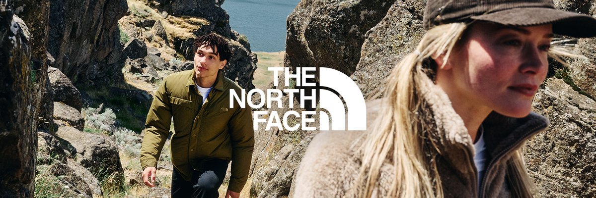 Two people navigating a rocky clifftop near open water, wearing The North Face jackets.