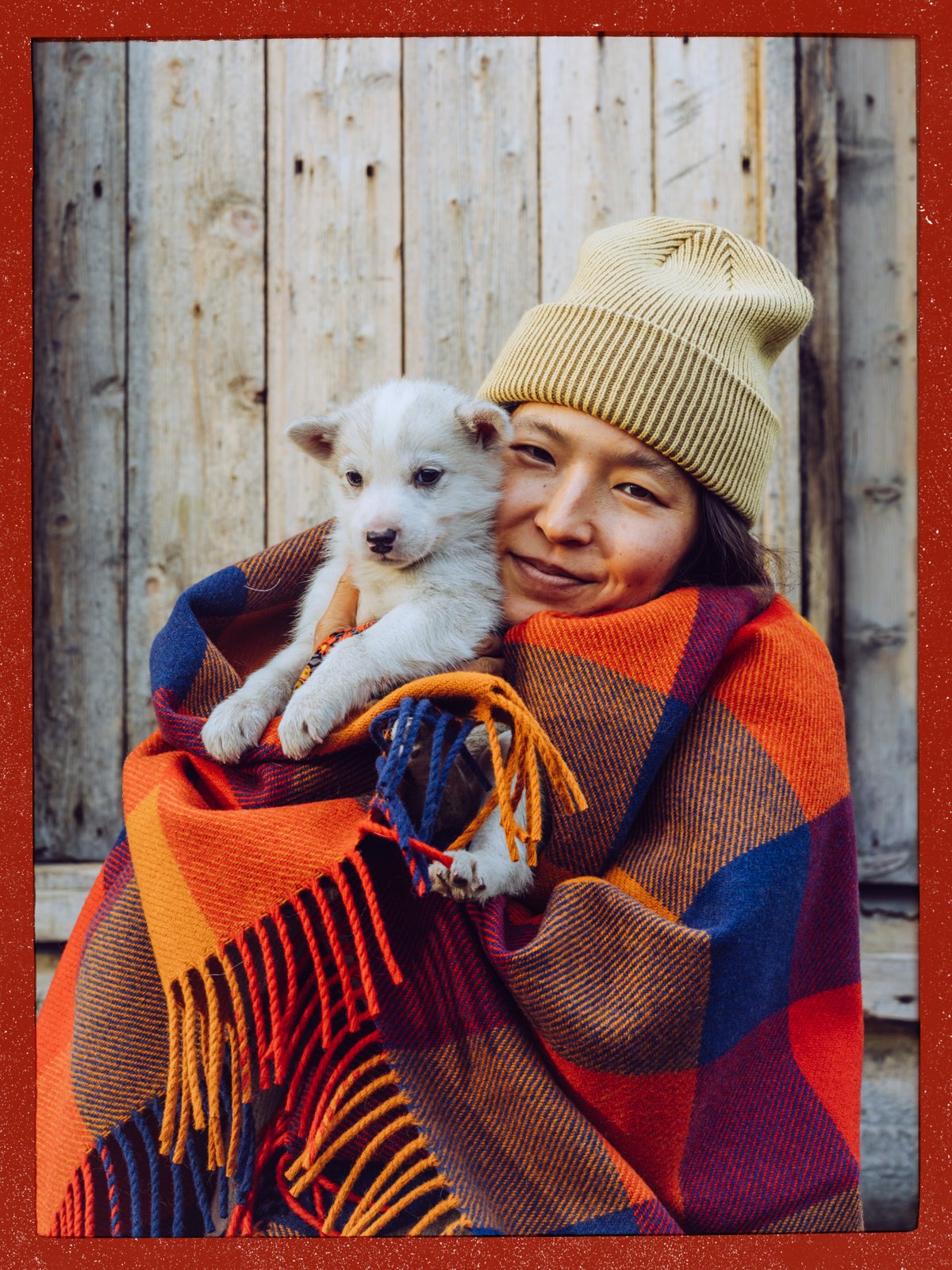 Person in a beanie wrapped in a plaid wool blanket, holding a white puppy against a weathered wood wall.