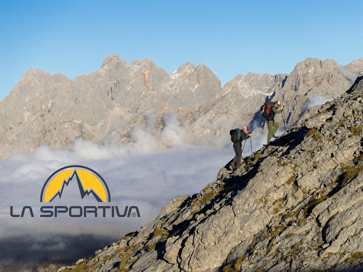 Two hikers using trekking poles to ascend a steep, rocky mountain ridge above the clouds; the La Sportiva mountain logo is in the bottom left.