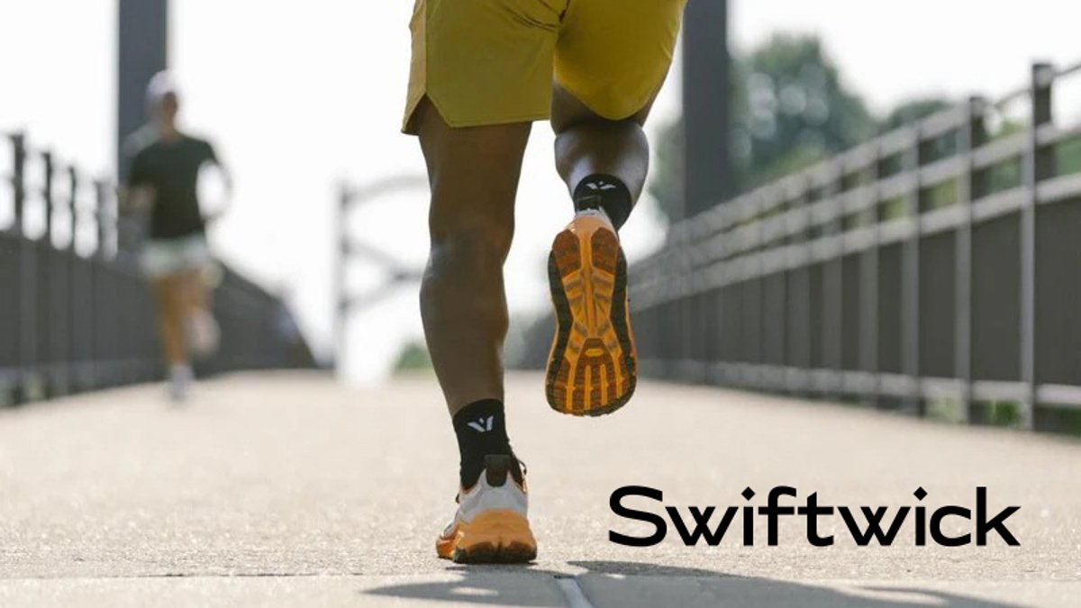 A close-up of a runner's foot and leg wearing yellow shorts and a running shoe with orange sole details on a paved path, with another jogger visible in the background.