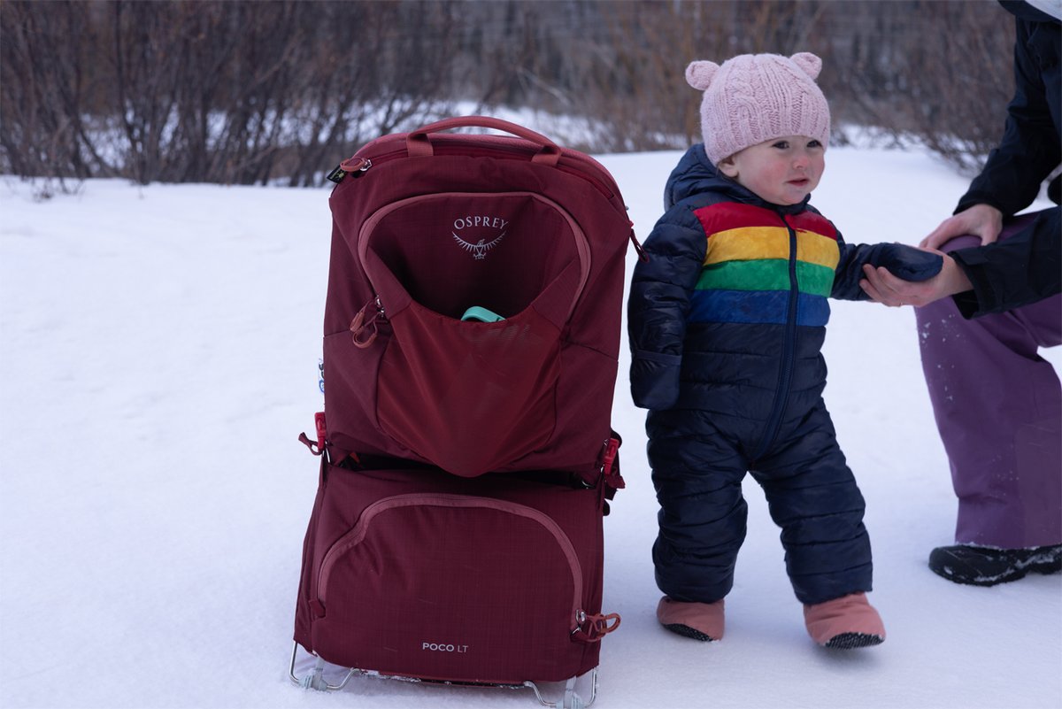 A toddler in a pink beanie stands near an Osprey Poco LT carrier in the snow.