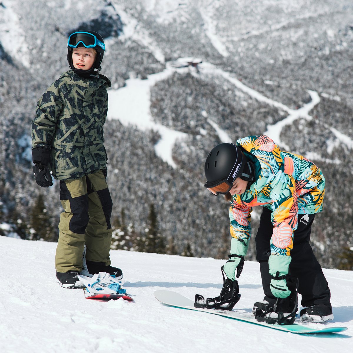 Two kids stand on top of a mountain. They are getting ready ot snowboard.