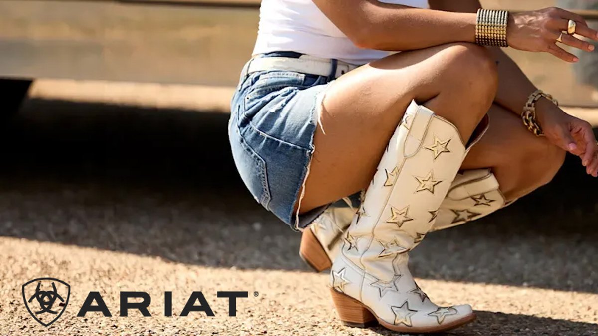 Person wearing cream-colored Ariat star-embellished cowboy boots with denim shorts and a white top, seated outdoors on sandy ground.