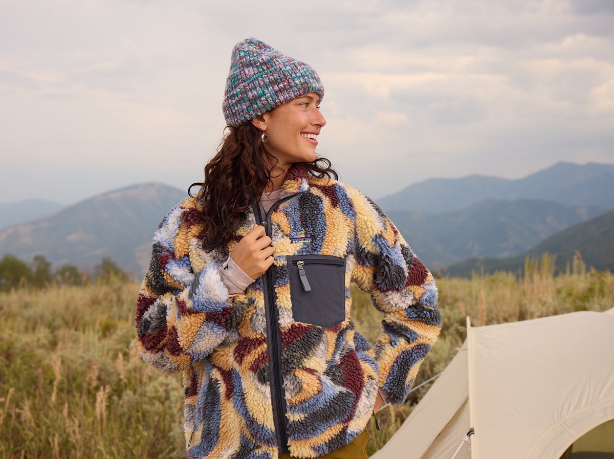 Woman wearing a patterned Patagonia sherpa fleece jacket and knit beanie, smiling in a mountain meadow at dusk next to her tent.