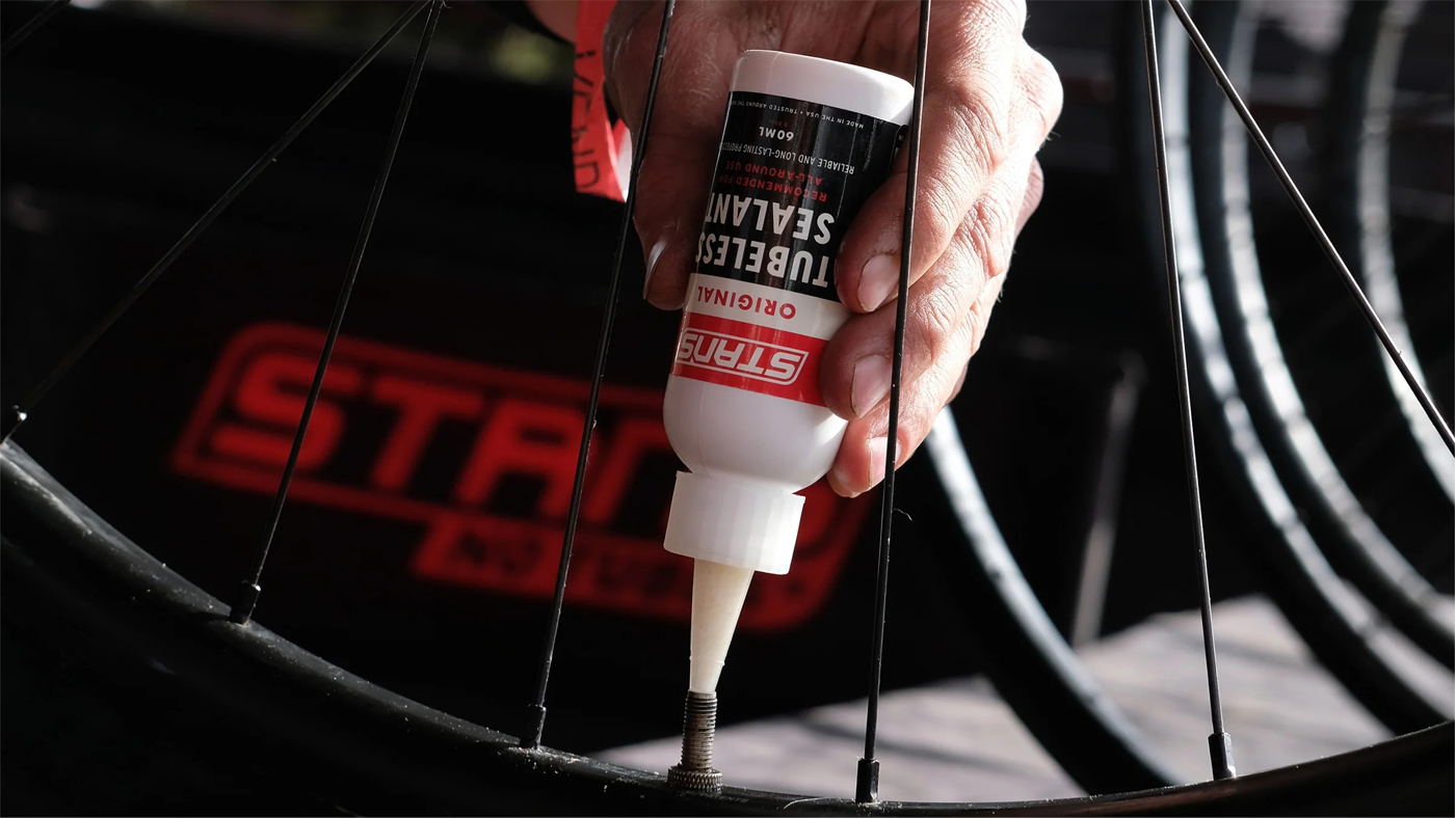 Person applying Stren bicycle tire sealant to a wheel rim with spokes visible in close-up detail.