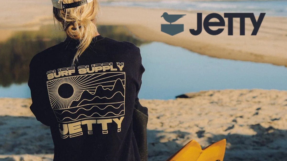 Woman wearing black Jetty Surf hoodie standing on sandy beach with surfboard and water view in golden hour sunlight.