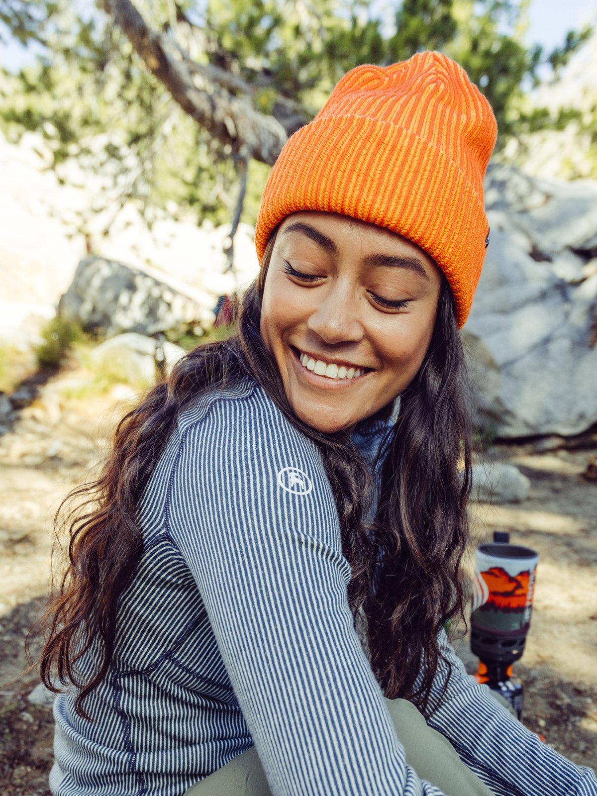 A woman with long black hair and a bright orange beanie hangs out at camp.
