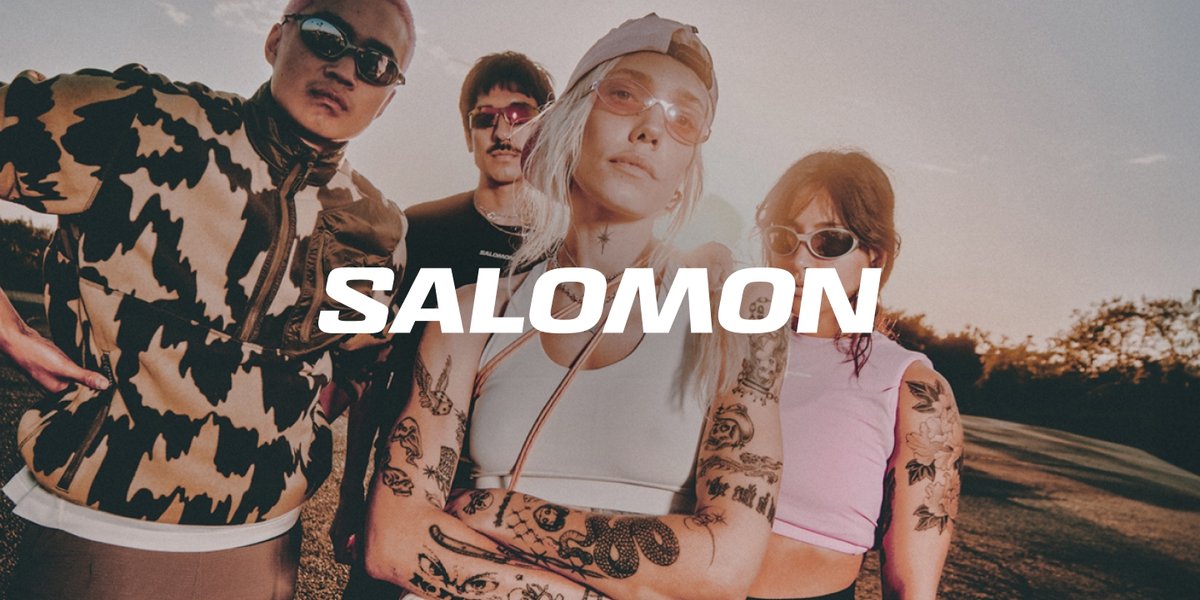 Two women with visible tattoos on their arms and two men stand outdoors on a hazy day. All four wear retro sunglasses and trendy running apparel. The word "Salomon" in bold white letters is centered across the image.