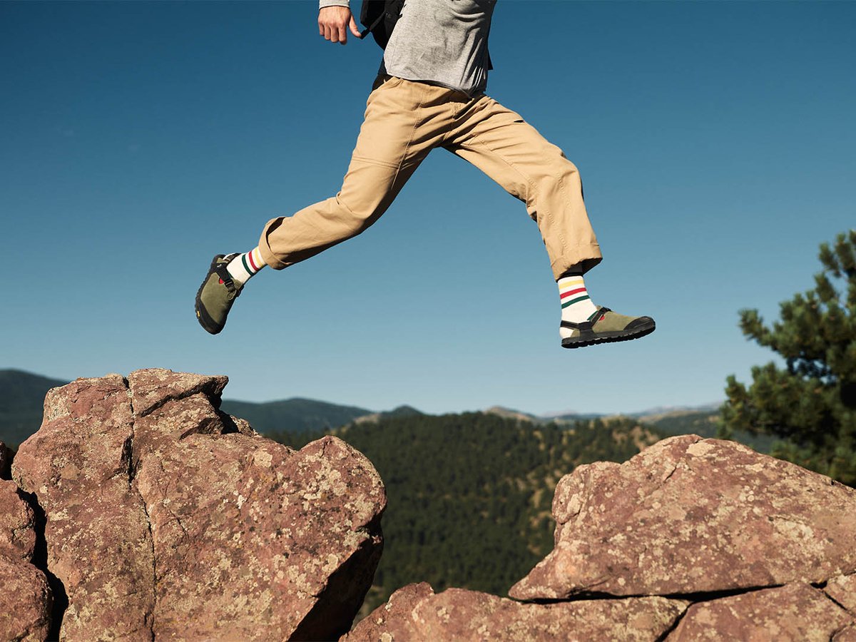 Person mid-jump between granite boulders in olive Bedrock Mountain Clogs and striped socks, with mountain ridgeline and blue sky behind