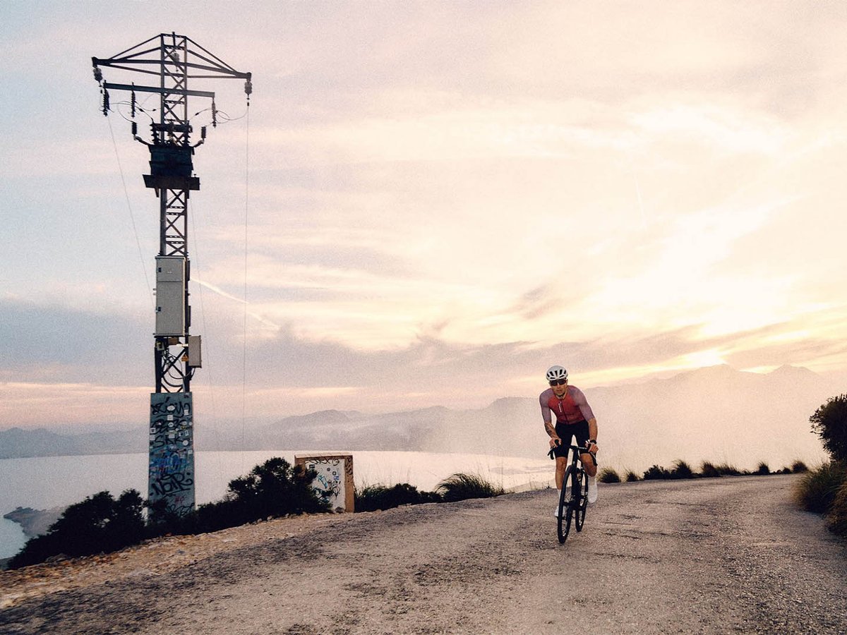 Road cyclist in a pink jersey and white helmet climbing a gravel-surface road past a graffiti-tagged electrical pylon, with a coastal mountain panorama and sunset sky behind.