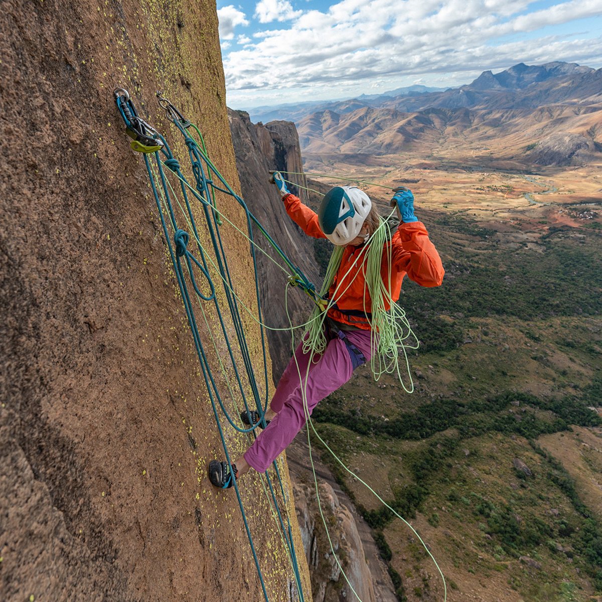 Climber wearing Patagonia climbing pants and helmet managing rope while hanging on steep granite big wall.