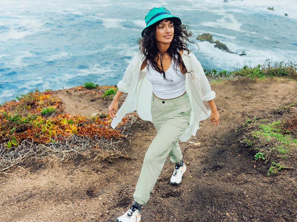 A woman walks on a trail near the ocean. Text overlay reads: Backcountry Wasatch Collection Durable Trailwear Shop Women’s.