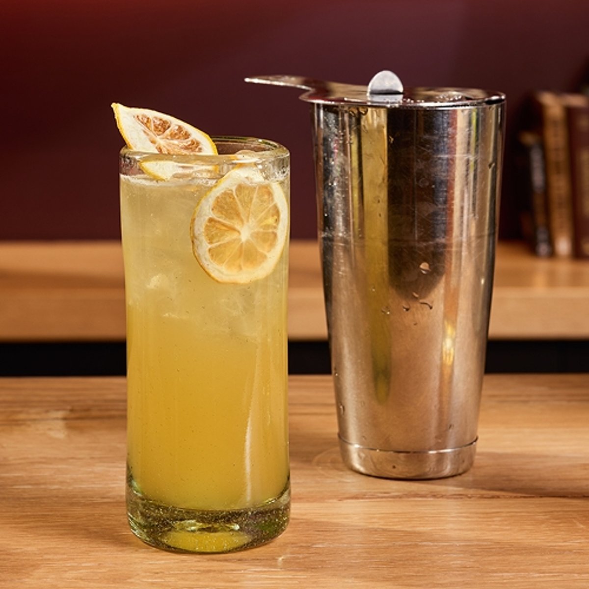 A bar countertop displays a tall cocktail garnished with dried lemon slices, a glass with more lemon slices, a bottle of High West Double Rye, a cocktail shaker, a jigger, wheat stalks, and books, all set against shelves filled with liquor bottles. Bold text at the top reads "Hopped up," with the High West Distillery and Backcountry logos.
