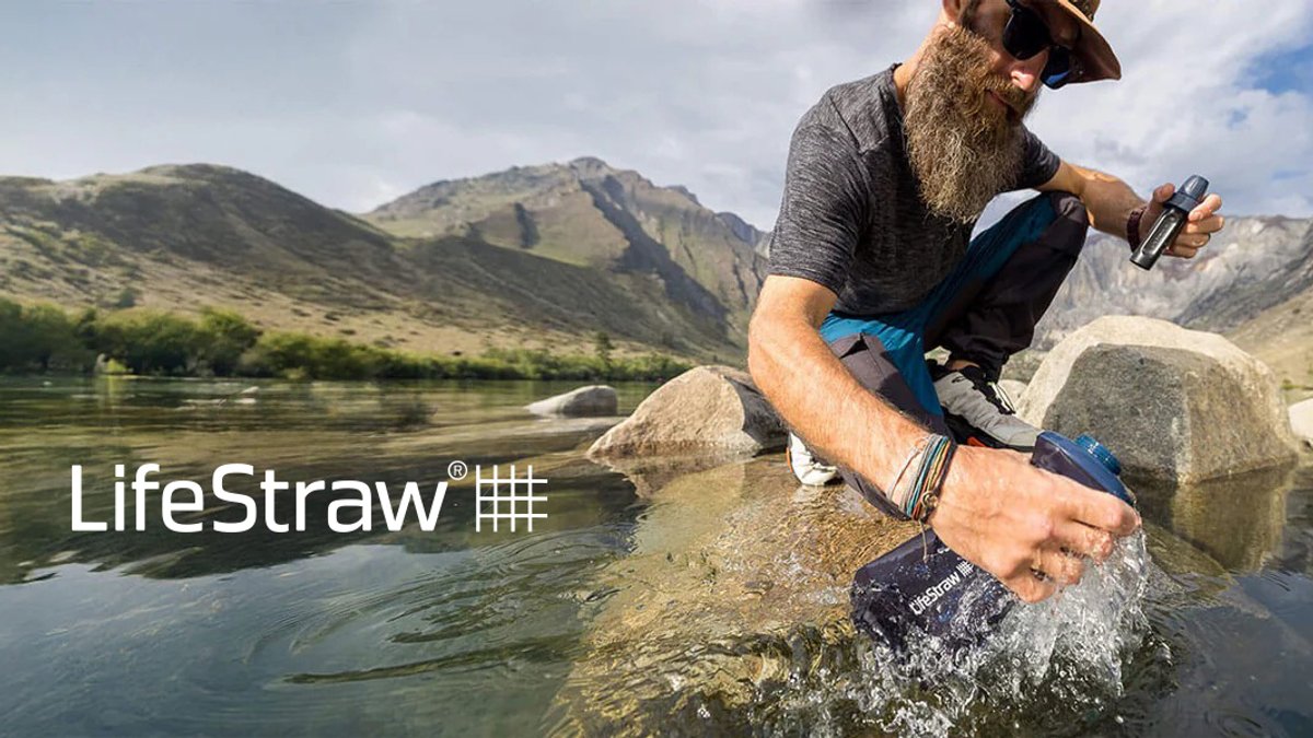 Bearded man using LifeStraw portable water filter while sitting on rocks by a mountain river during outdoor adventure.
