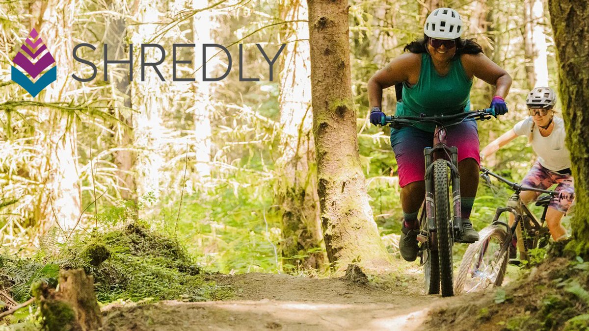 Two female mountain bikers ride together on a forested trail, showcasing SHREDLY outdoor adventure gear and cycling equipment in a natural woodland setting.