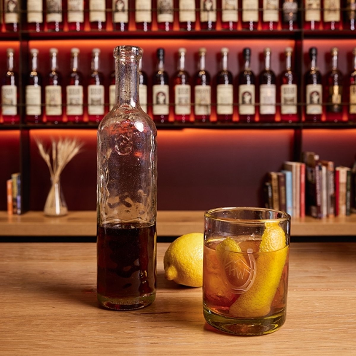 A bar countertop displays ingredients for a cocktail, including a bottle of High West Campfire whiskey, a bottle of Angostura aromatic bitters, a lemon, a mixing spoon, and a glass filled with an old fashioned cocktail, with a background of shelves lined with liquor bottles. The top of the image has the text “Campfire Old Fashioned” below the High West Distillery and Backcountry logos.