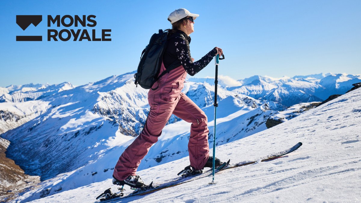 A skier in burgundy pants and a white cap ascends a snowy mountain slope with a trekking pole, with snow-covered peaks visible in the distant background.