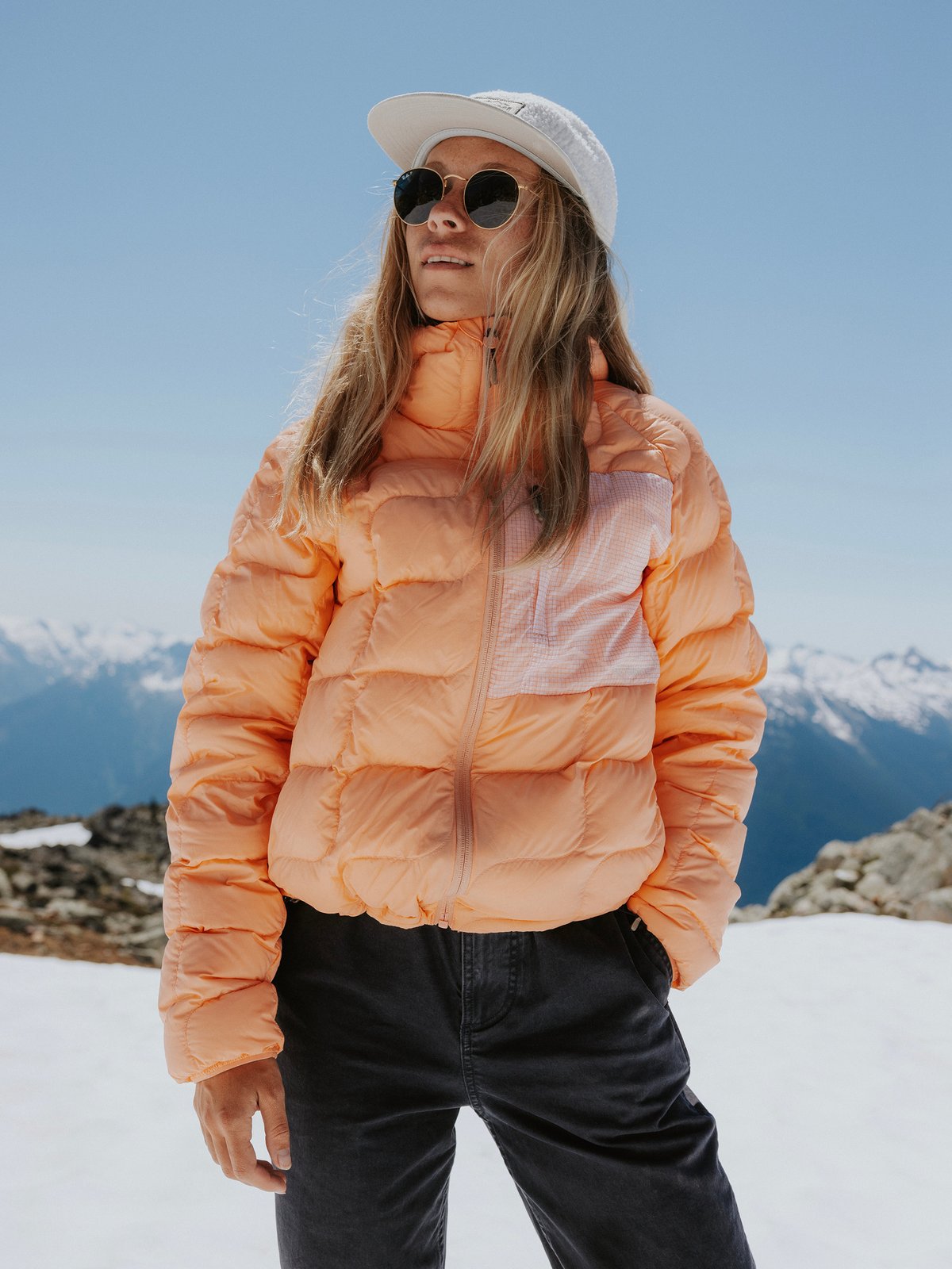 A woman with long hair wearing a green quilted jacket, white sweater, and brown pants stands in a pine forest.