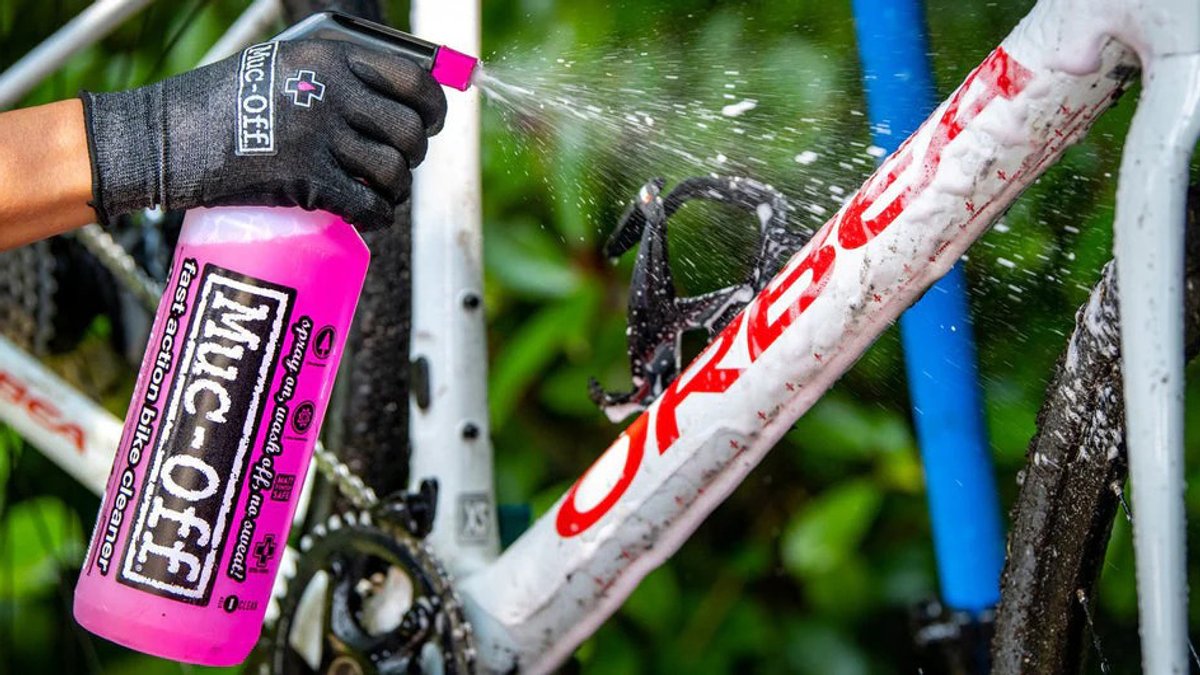 A person wearing work gloves sprays pink bike cleaning product onto a mountain bike frame with red and blue components.