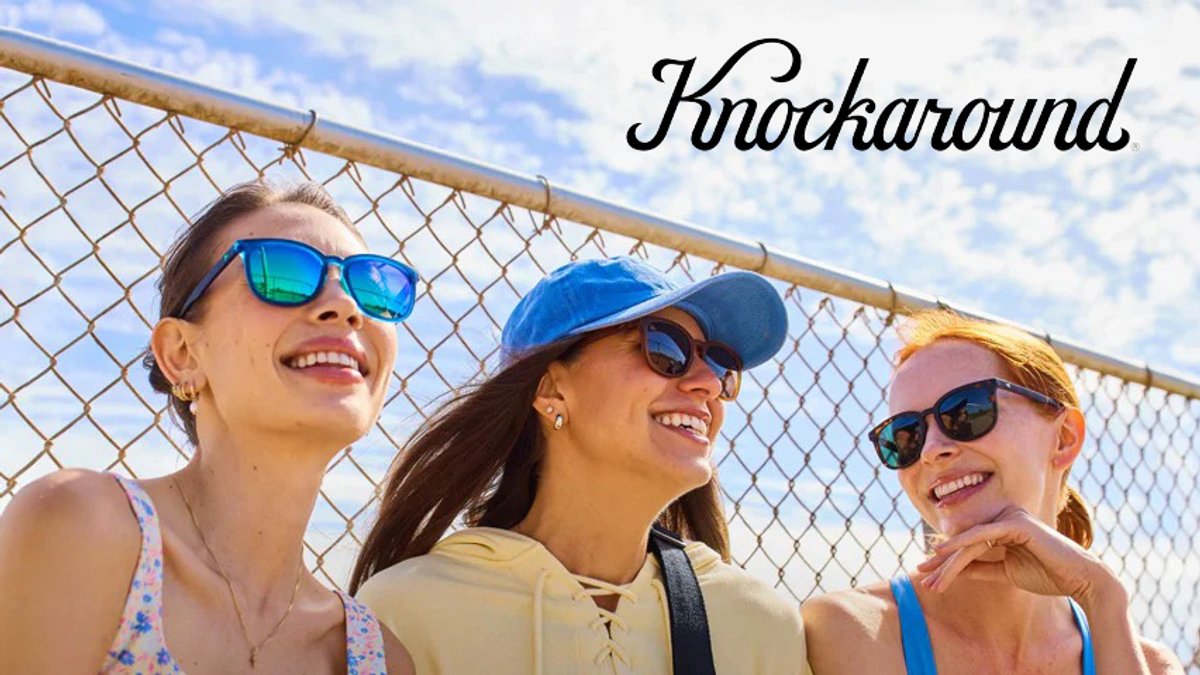 Three smiling women wearing blue-tinted Knockaround sunglasses and casual summer apparel stand together against a chain-link fence on a sunny day, showcasing outdoor eyewear and vacation style.