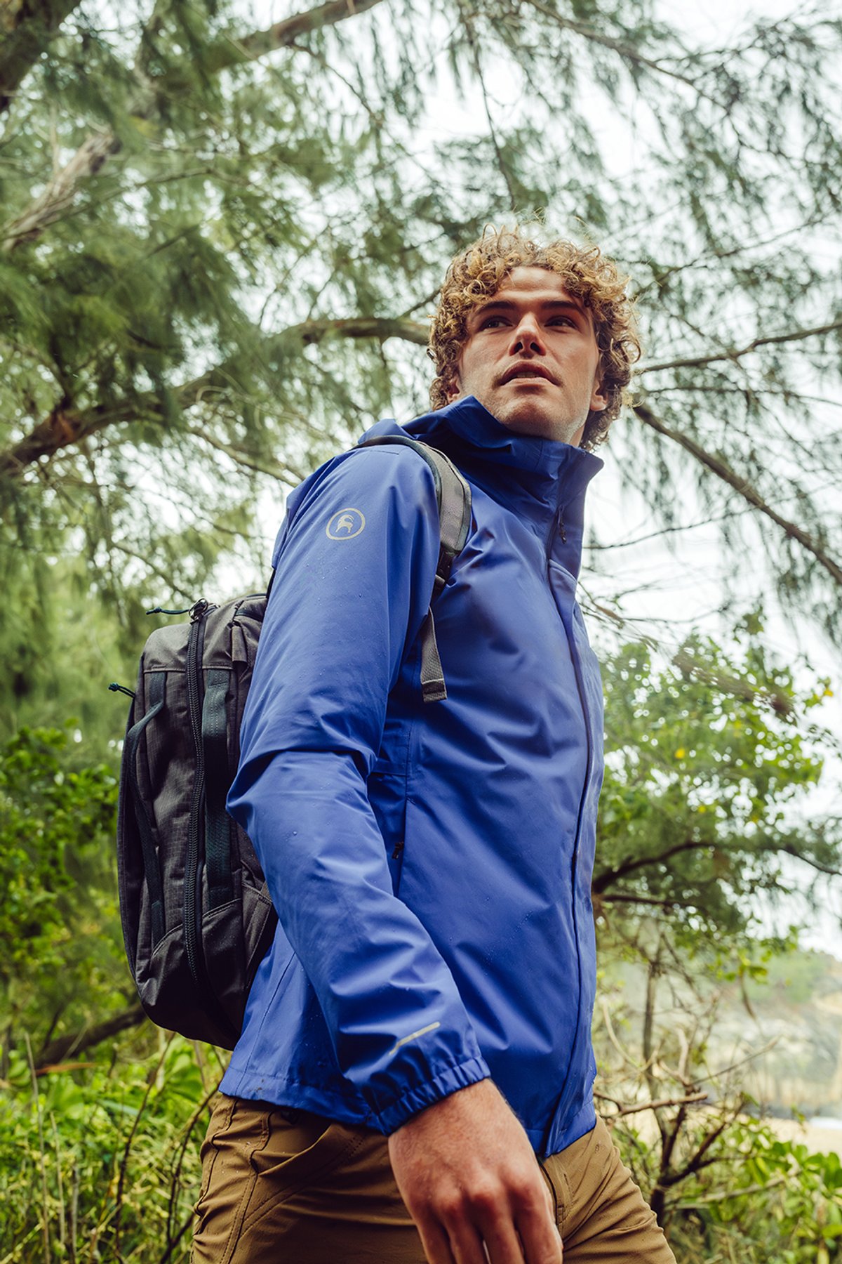 Man wearing blue Backcountry hooded rain jacket with backpack hiking through wet tropical forest terrain