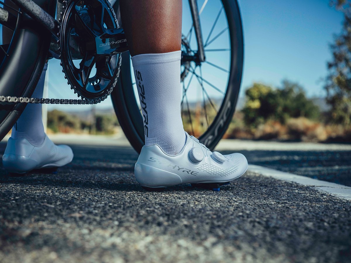 Rear view of a white S-PHYRE shoe standing beside a bike, low ground angle, Dura-Ace crank and S-PHYRE sock lettering visible in the background
