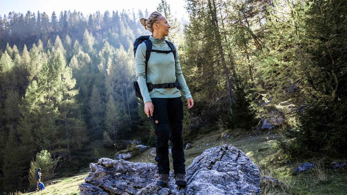 A woman wearing the Fjallraven Keb Agile pants stands on a rock against a forest landscape.