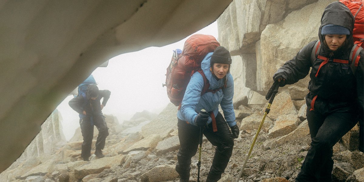 Three backpackers walk through the rain, taking shelter under a rocky overhang. 