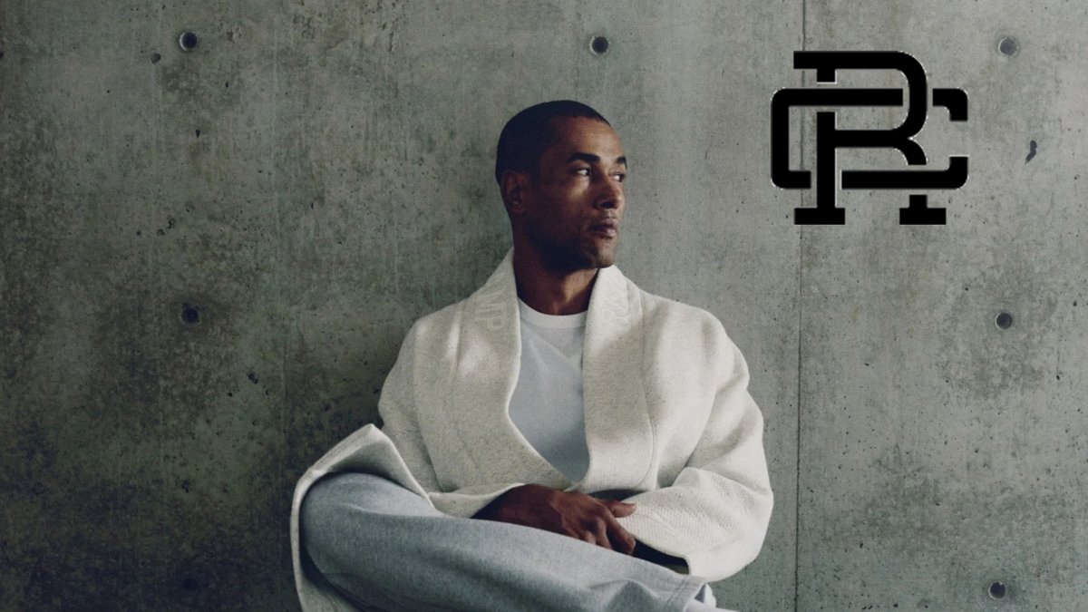 A man wearing a cream-colored cardigan and white t-shirt sits against a concrete wall, looking thoughtfully to the side.