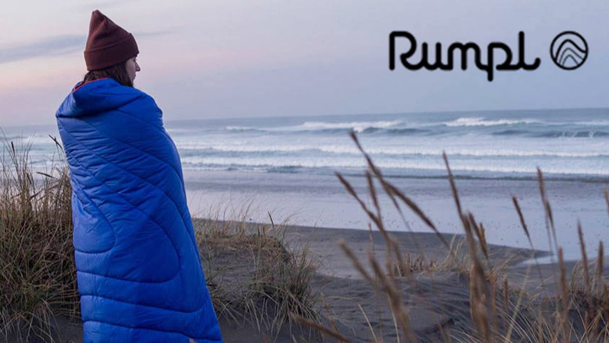 Person wrapped in a blue Rumpl quilted camping blanket standing on a beach at dusk overlooking ocean waves and coastal dunes.