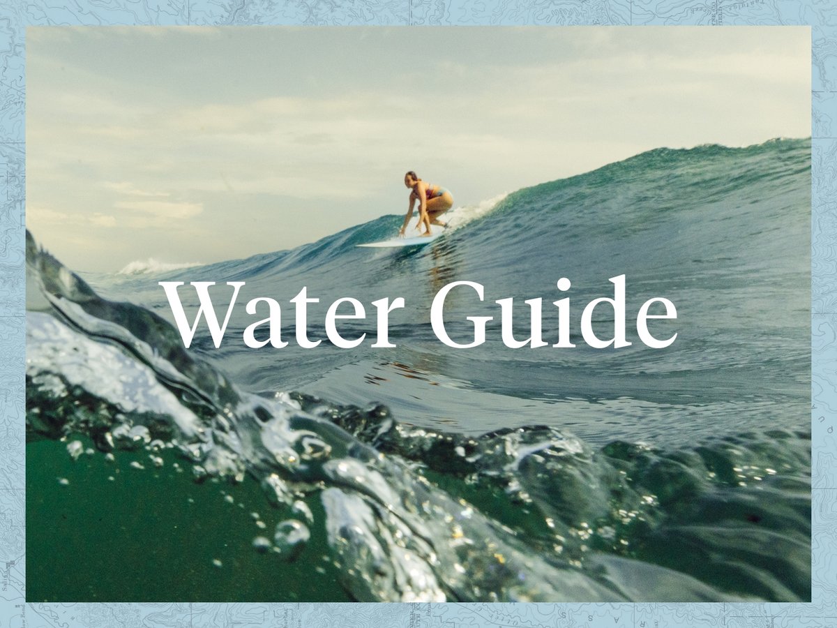 A surfer begins to stand on their board. The camera is slightly submerged in the water. The words read, “Water Guide”.