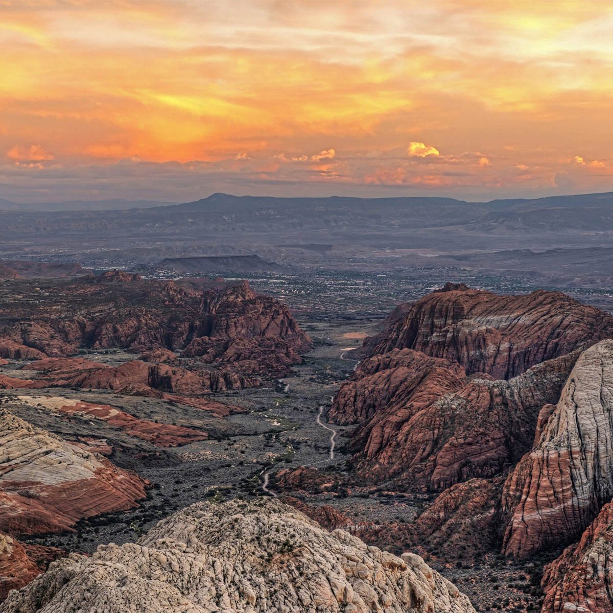 Elevated view across Snow Canyon's red and white striped sandstone formations toward a valley and distant mountain range under an orange sunset sky.