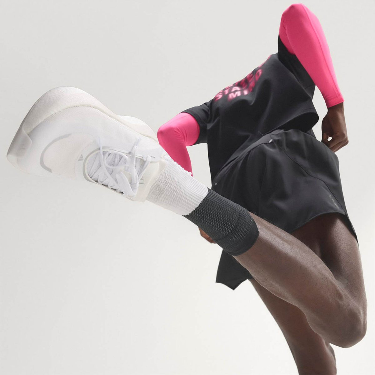 Runner with knee driven high toward camera, showing the bottom of a white Adidas running shoe with off-white midsole, wearing black shorts and pink arm sleeves, on a white background.