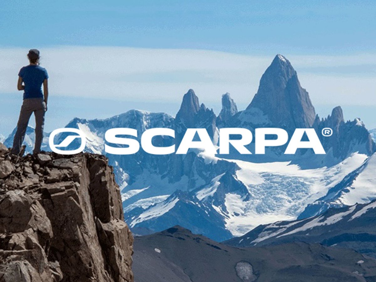 Scenic mountain landscape with a person standing on a rocky cliff overlooking snow-covered peaks and a dramatic alpine spire, with the SCARPA logo displayed across the center.