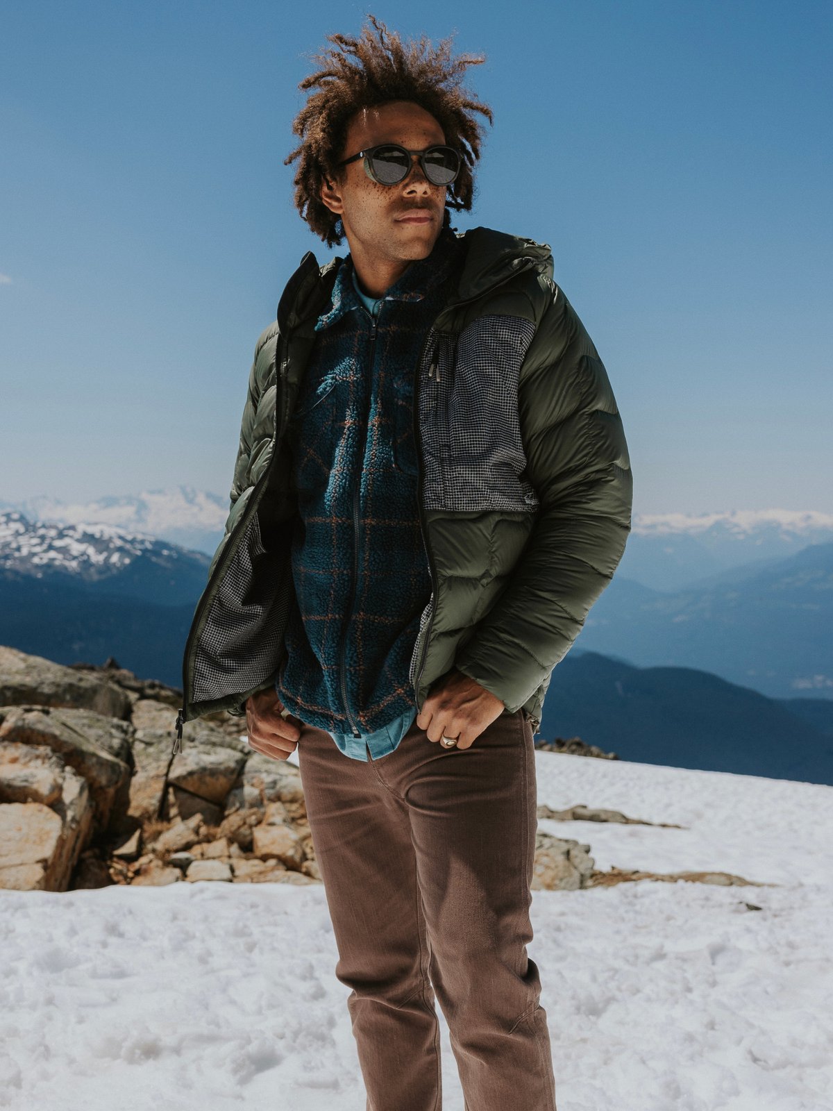 A man on a mountaintop wearing a Backcountry Waas ALLIED Down puffy jacket, plaid fleece, and round glacier glasses. 