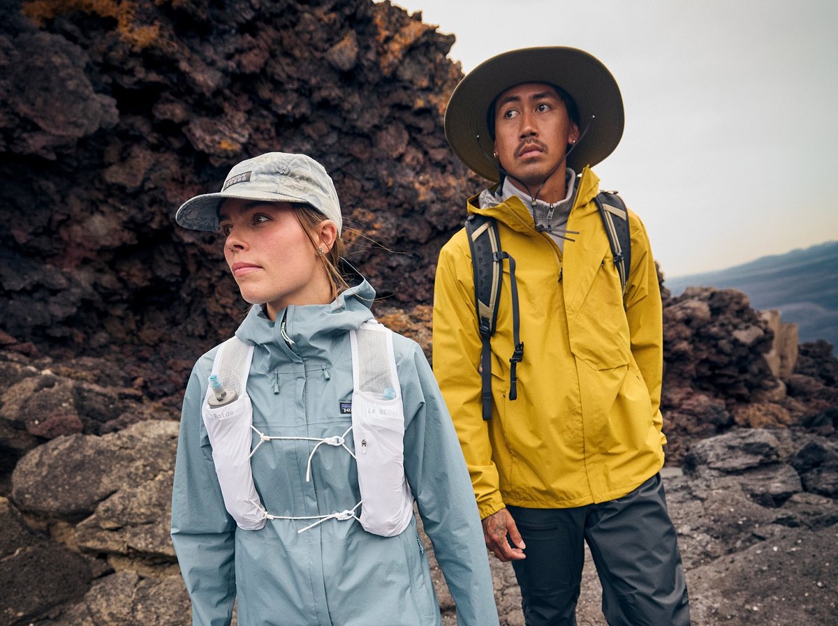 Two hikers navigate rocky terrain in technical outdoor gear — blue and yellow jackets with backpacks against mountain landscape.