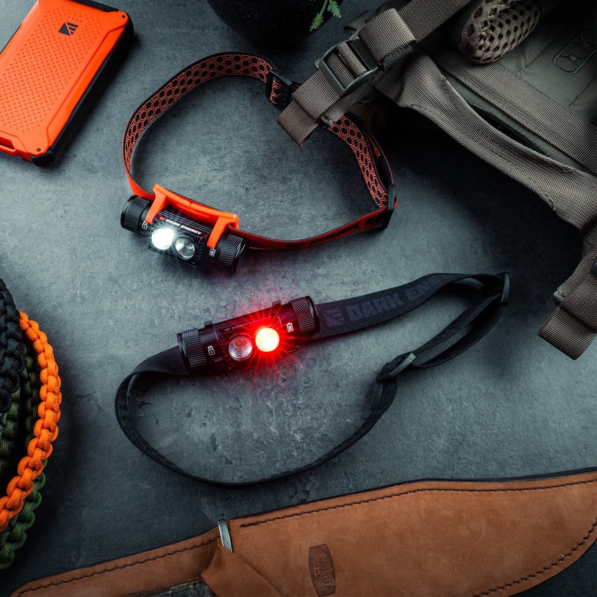 Two Dark Energy Oracle headlamps on a slate surface, one lit white and one lit red, next to an orange Poseidon power bank, coiled paracord, and a pack.