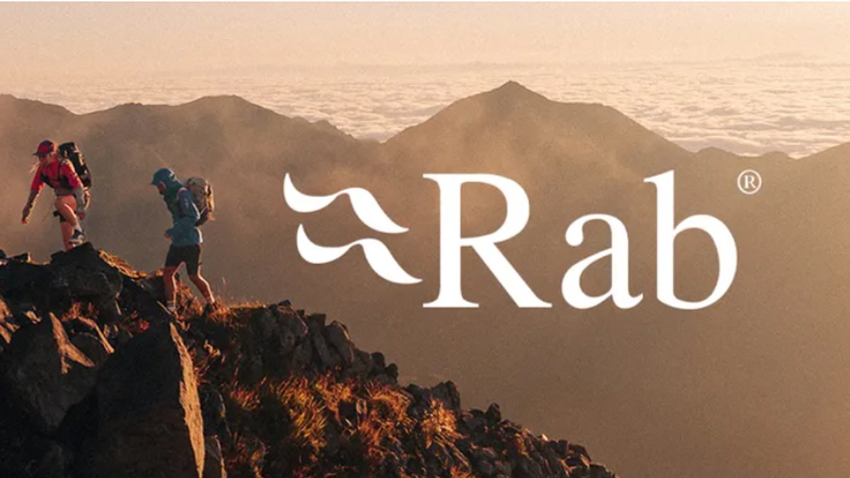 Two hikers scramble up a jagged, rocky mountain ridge at sunset, with a sea of clouds filling the valley behind them and the white Rab logo centered.
