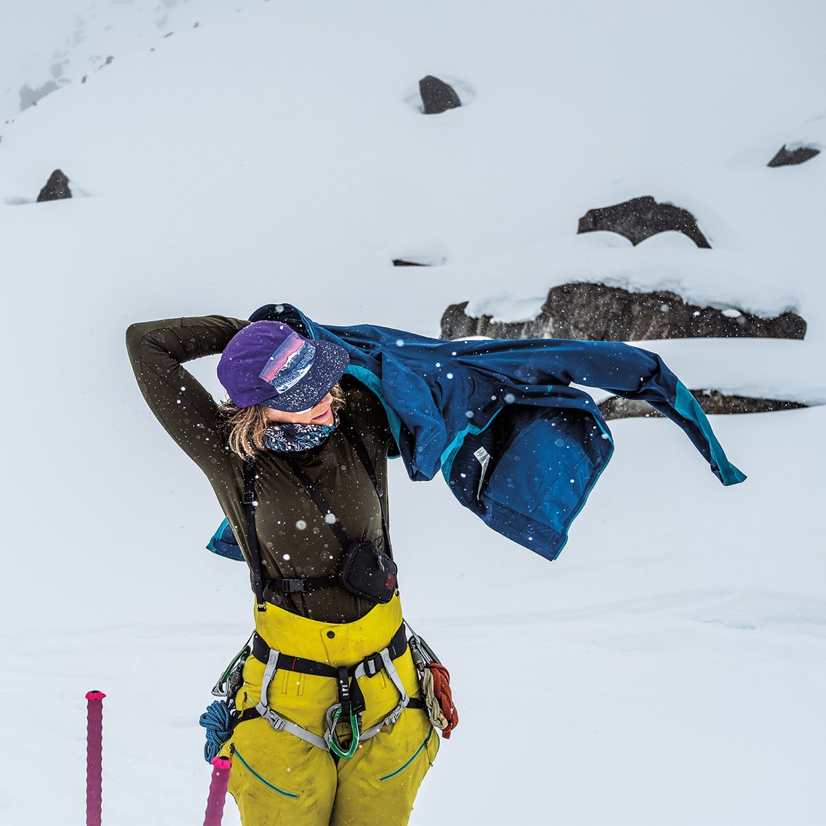 Woman in an olive green Capilene baselayer top pulls on a blue jacket over her climbing harness during a snowy whiteout on a glacier.
