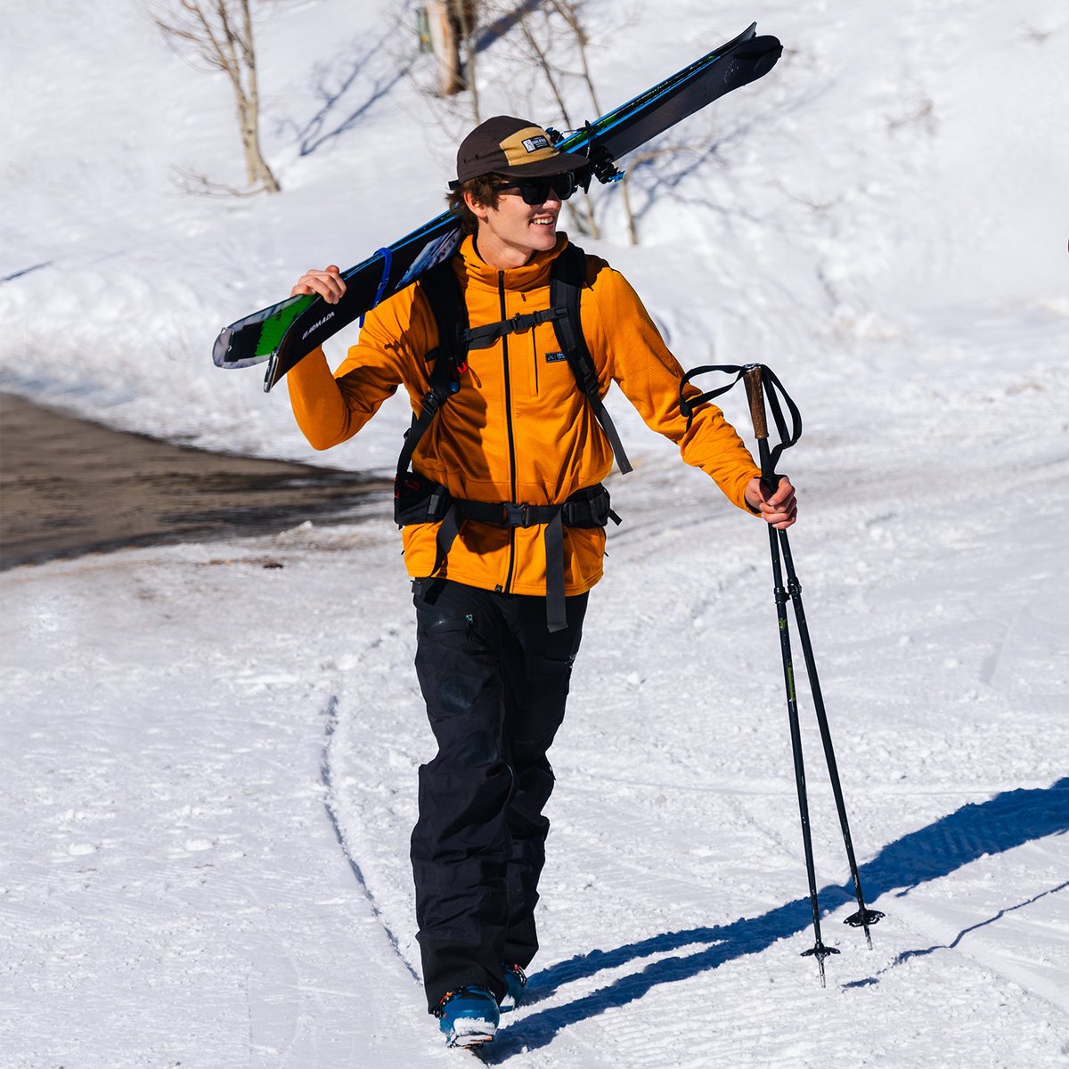Backcountry skiier wearing Coalatree workwear hat walking  in ski boots and carrying skis on snowy road.