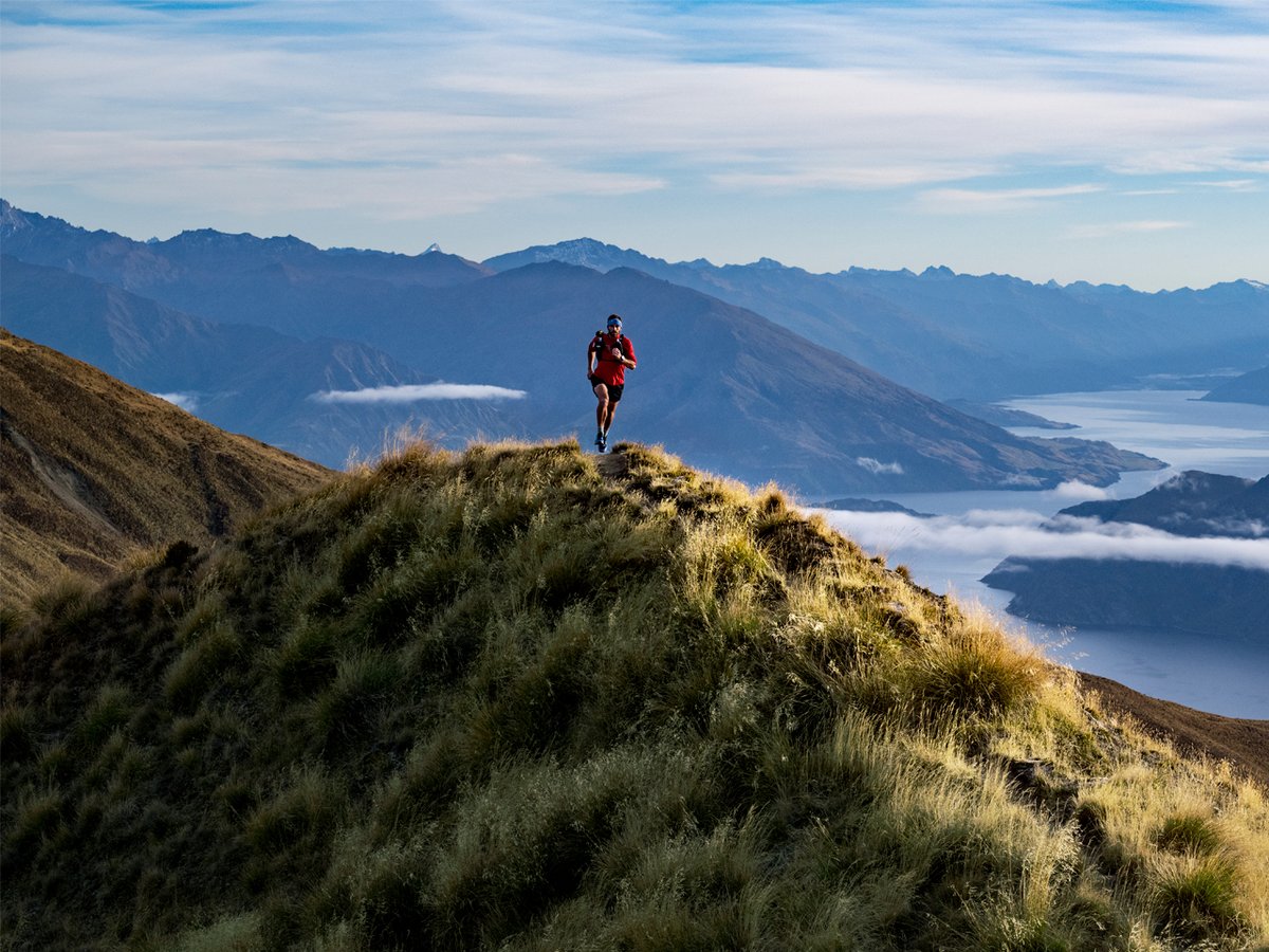 A person runs along a mountain peak, a panoramic view of a foggy mountain range behind them.