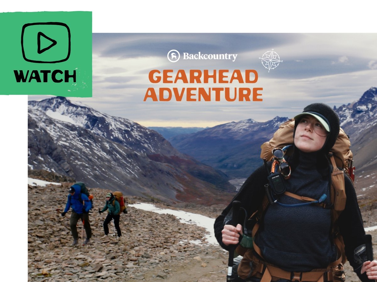 Three hikers with large backpacks and trekking poles ascend a rocky mountain trail. Snow-capped peaks and a dramatic cloudy sky fill the background. A flag in the upper left corner shows an illustrated play button and includes the text “Watch”.