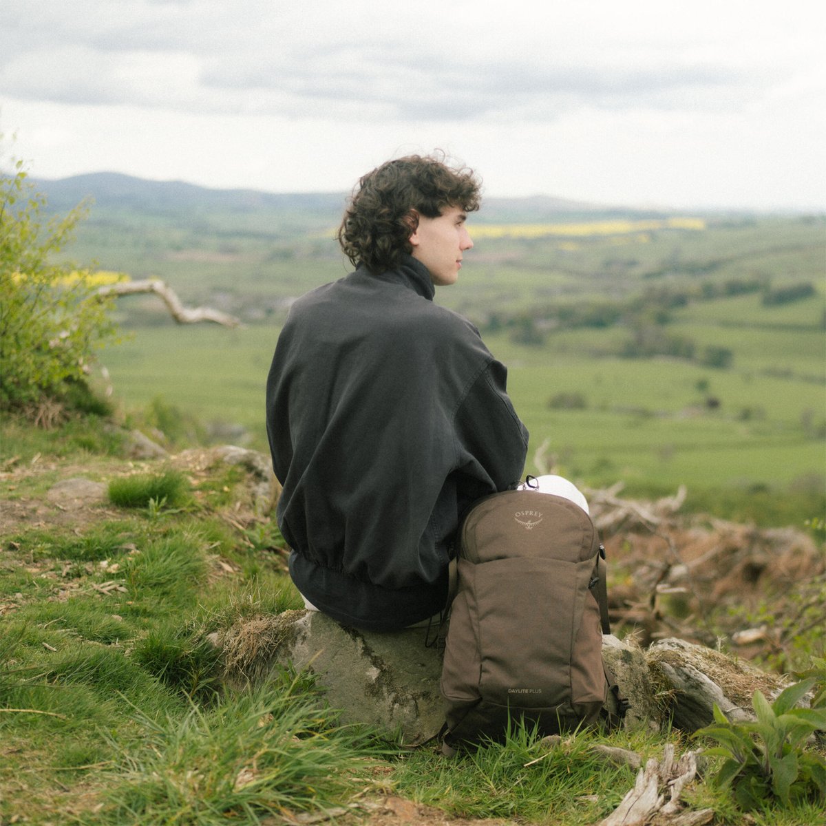 A person sits outside in a green field, an Osprey Daylite pack on the ground next to them 