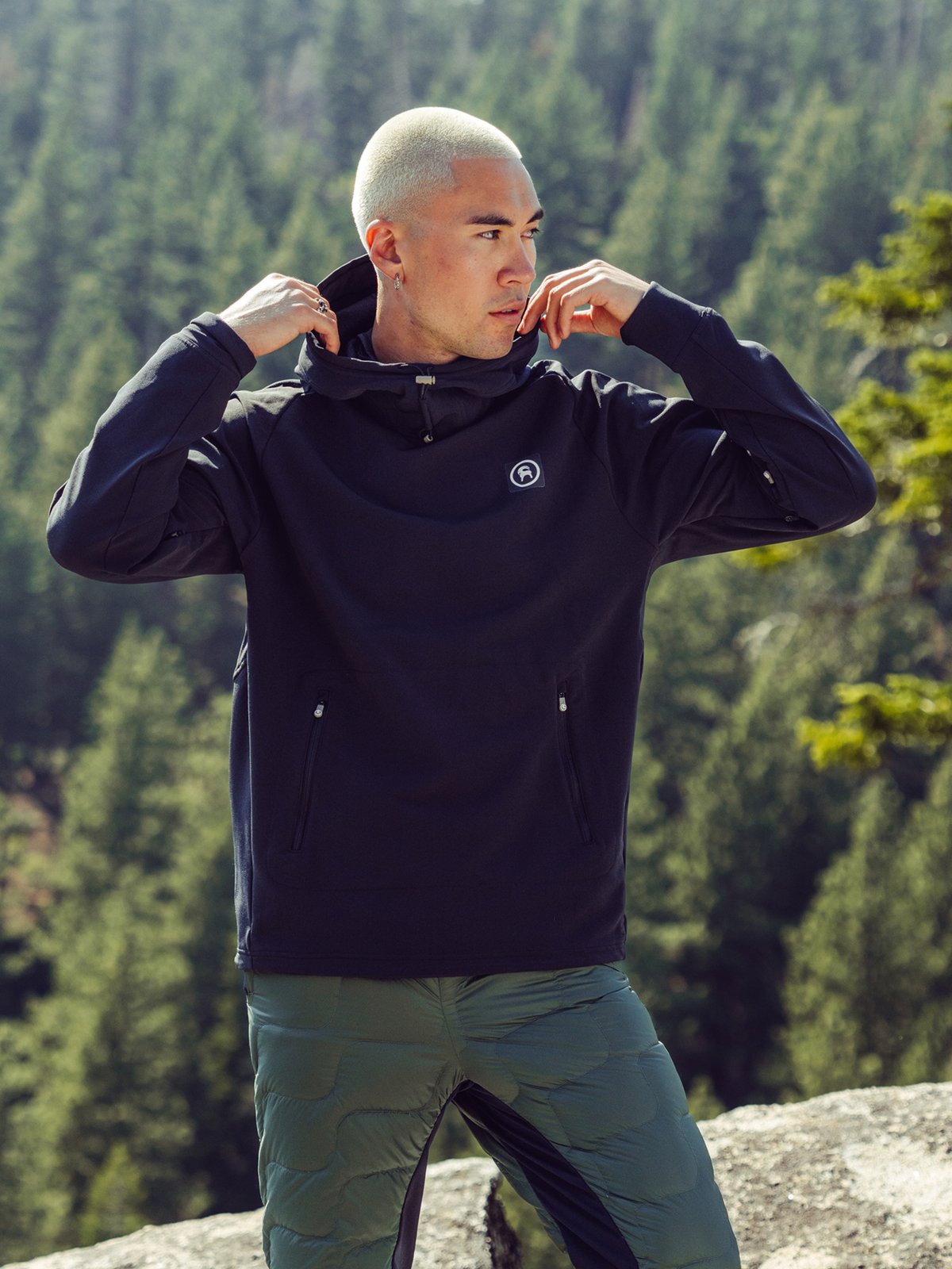 A man in a forest adjusts the collar of a black pullover with a small circular goat logo on the chest.