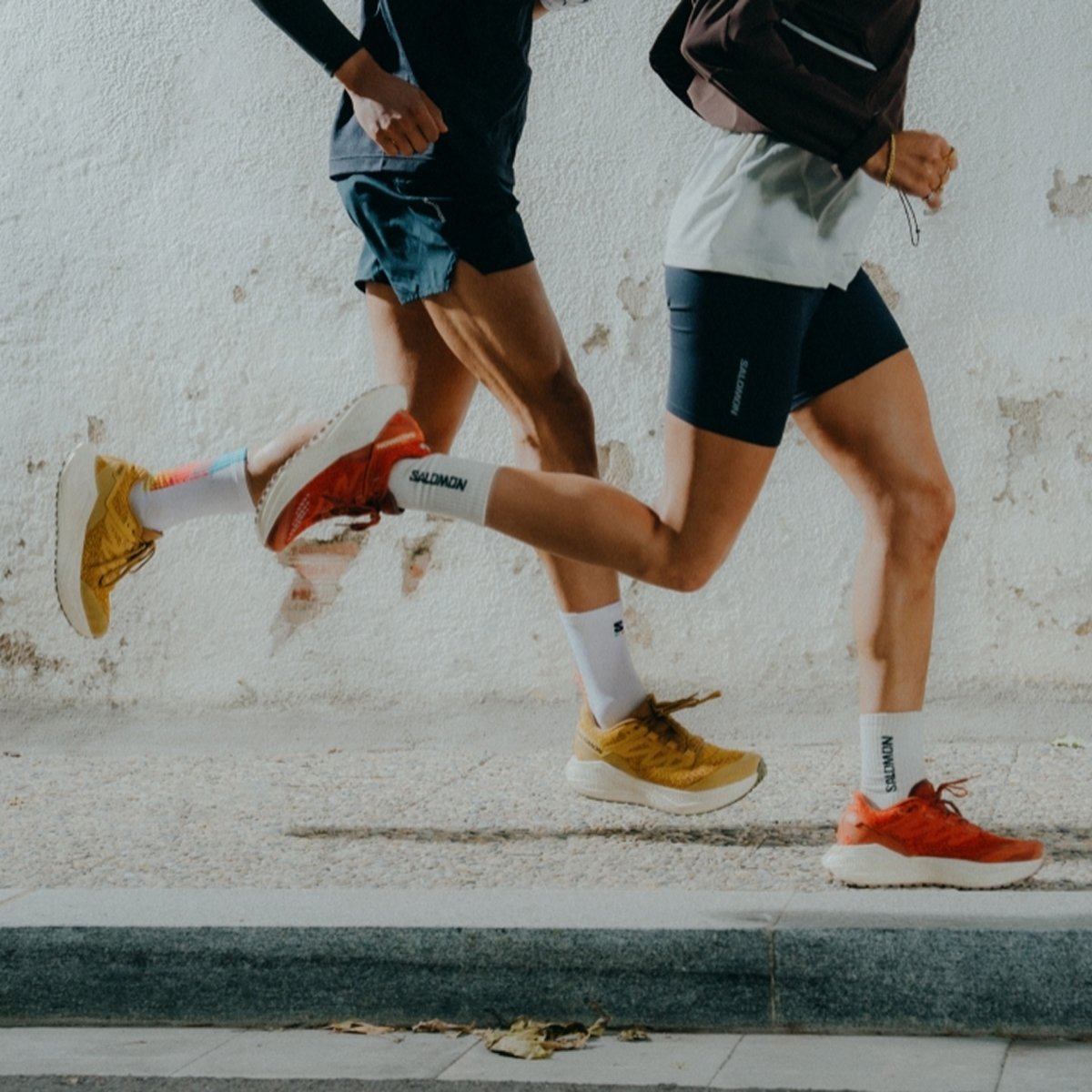 Two people in running attire are jogging side by side on a sidewalk, wearing Salomon brand sneakers—one pair yellow and the other orange—with white Salomon socks visible above their shoes.