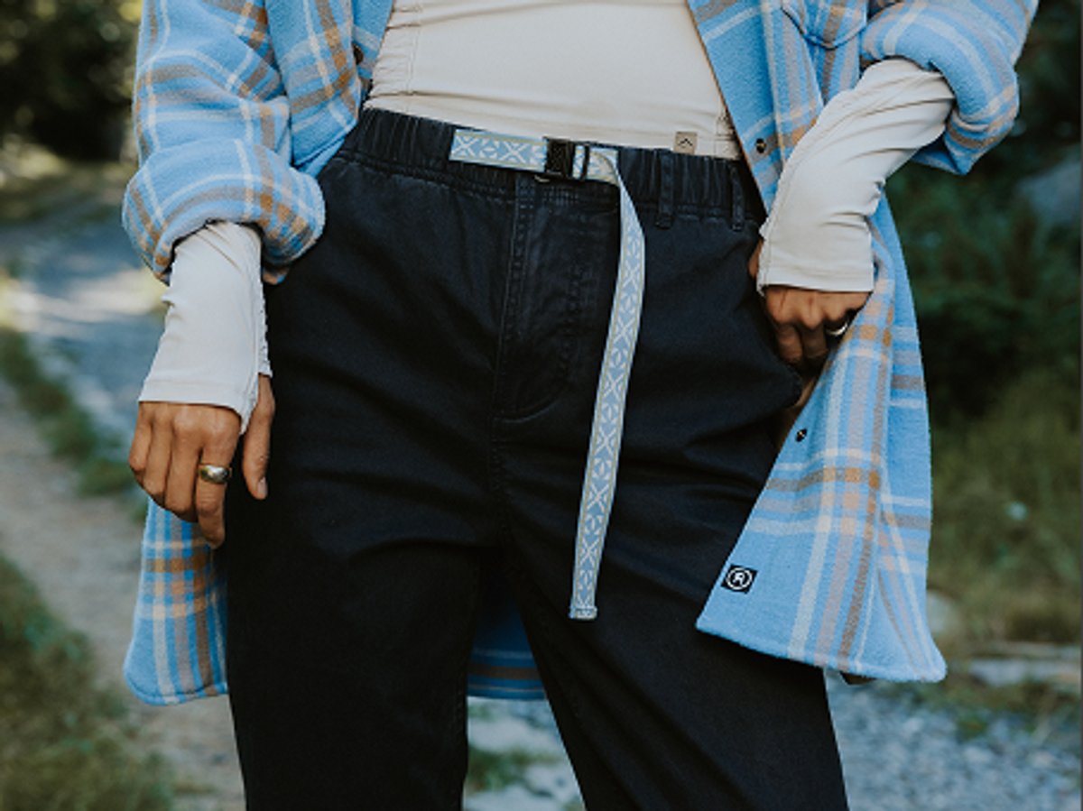 Close-up of a person wearing black pants with a woven belt, layered with a long-sleeve top and plaid shirt in an outdoor setting.