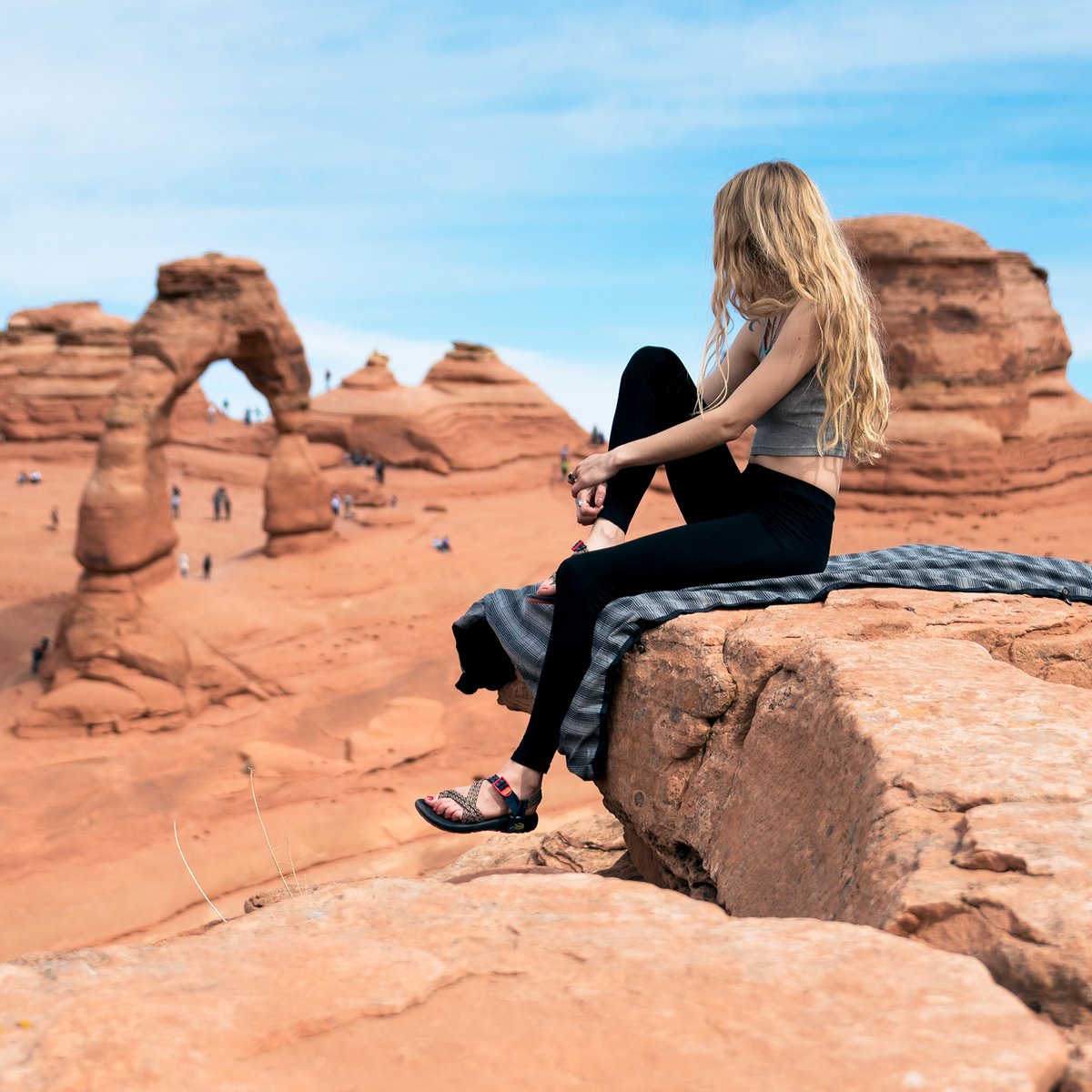 Person sitting on Coalatreee Katchula blanket on sandstone rock overlooking Delicate Arch in Moab, Utah.