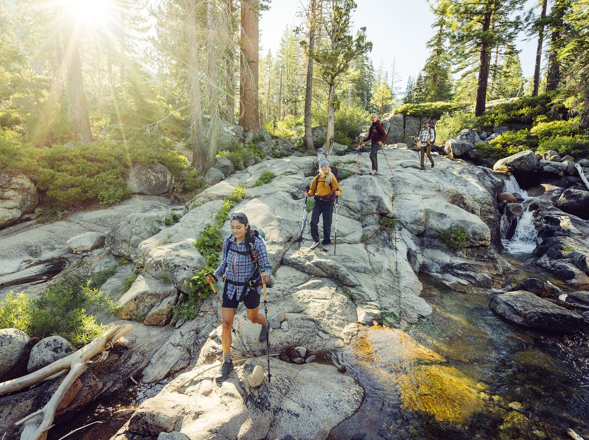 Four hikers with trekking poles pick their way across sun-warmed granite slabs beside a cascading Sierra Nevada creek, tall pines overhead.