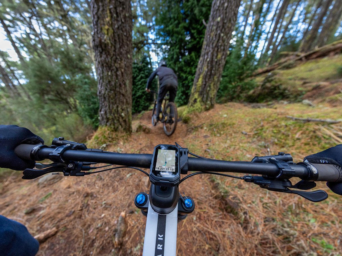 Mountain biker’s cockpit view chasing a rider down a narrow forest singletrack, with a Garmin bike computer mounted on the handlebar showing the route.